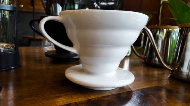 Beyaz çömlekli kahve fincanı, mermer kaplı ahşap zemin, cam ve gümüş parlayan mutfak malzemeleriyle çevrili.