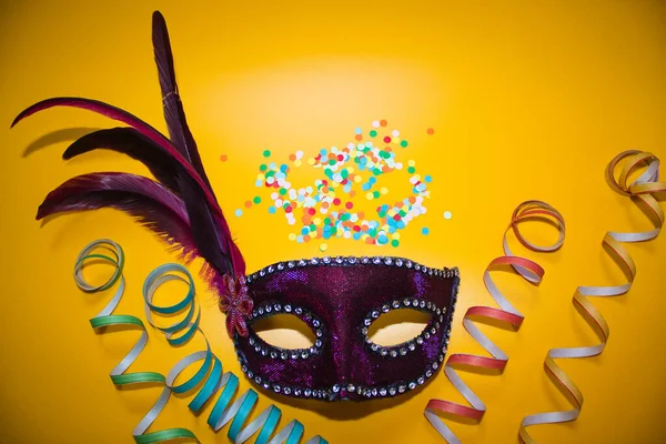 Sarı arka planda konfeti ve kıvırcık kağıtlı mor karnaval maskesi.