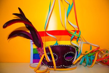 Sarı arka planda konfeti ve kıvırcık kağıt ile mor karnaval maskesi ve gay gurur bayrağı