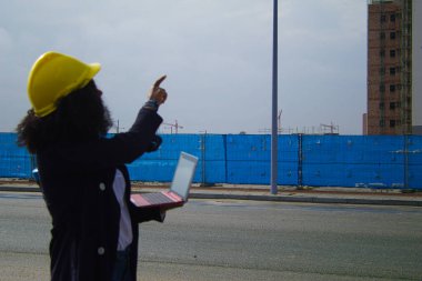 African-American black woman construction manager in yellow hardhat set out to check the construction of the flats. She is holding a computer in her hand to check the progress of the construction