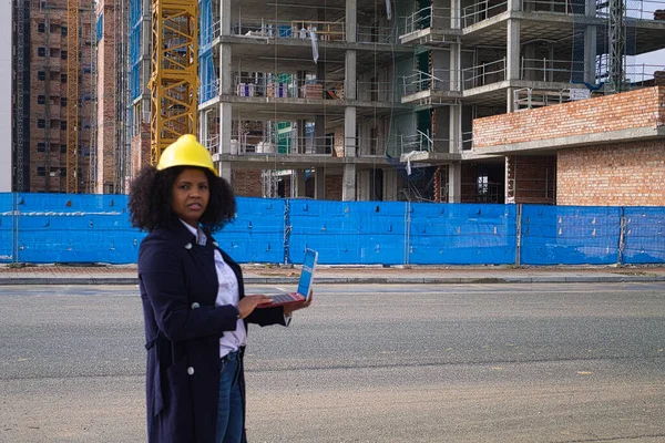 African-American black woman construction manager in yellow hardhat set out to check the construction of the flats. She is holding a laptop in her hand to check the progress of the construction