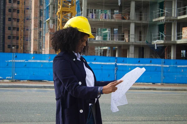 African-American black woman construction manager in yellow hardhat sets out to check the construction of the flats. she is holding plans in her hands and is looking at the progress.