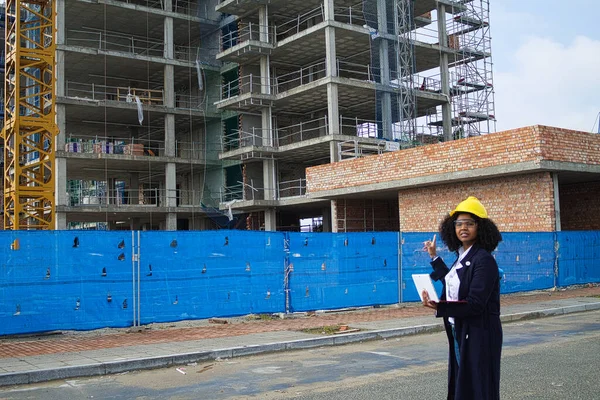 African-American black woman construction manager in yellow hardhat sets out to check the construction of the flats. She is holding a computer in her hand to check the progress of the construction