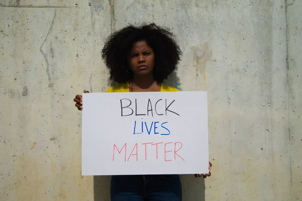 angry-faced african-american woman holds up a banner reading black lives matter. In the background grey wall