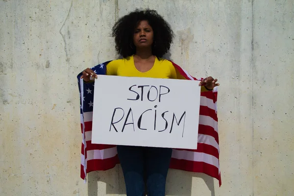 african-american woman holds a stop racism banner in her hands and united states flag over her shoulders. In background grey wall.