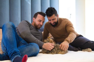 Yatakta oturmuş evcil hayvanlarıyla oynayan eşcinsel bir çift. Köpeğe sevgiyle bakıyorlar.