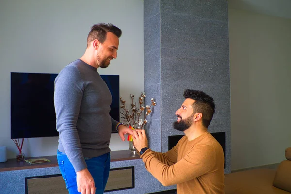 homosexual man kneeling down and asking his partner to marry him in the living room of his house. In the background you can see the gay pride flag