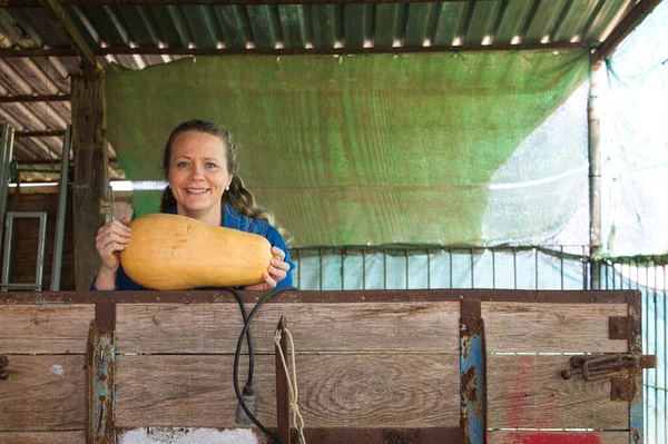 pretty blonde farm woman on a work trailer showing part of her harvest. It is a long orange pumpkin