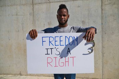 Afro-Amerikalı bir adam elinde özgürlüğün bir hak olduğunu gösteren bir pankart tutuyor. Adamın sol elinde kelepçe ya da kelepçe var. İnsan hakları. Irkçılığı bırak. Siyah hayatlar önemlidir..