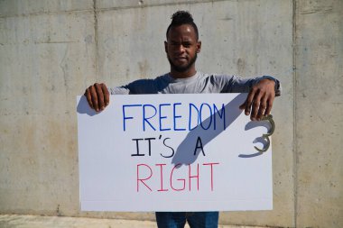 Afro-Amerikalı bir adam elinde özgürlüğün bir hak olduğunu gösteren bir pankart tutuyor. Adamın sol elinde kelepçe ya da kelepçe var. İnsan hakları. Irkçılığı bırak. Siyah hayatlar önemlidir..