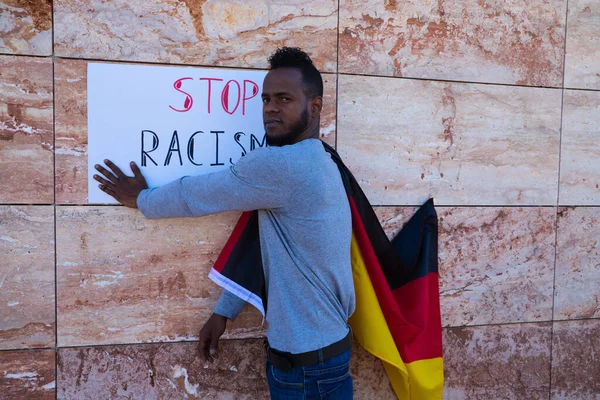An African-american man on a wall holds a sign that reads stop racism. The man is carrying a German flag on his shoulders.