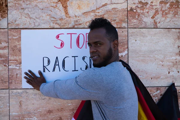 An African-american man on a wall holds a sign that reads stop racism. The man is carrying a German flag on his shoulders.