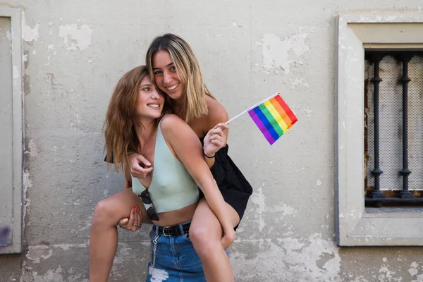 two beautiful young women on holiday. The women are lesbians and one is carrying the other. She carries a gay pride flag in her hand and they are a couple. Concept of equality and diversity.