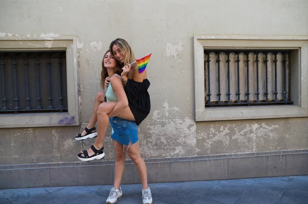 two beautiful young women on holiday. The women are lesbians and one is carrying the other. She carries a gay pride flag in her hand and they are a couple. Concept of equality and diversity.