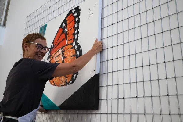 Olgun kadın ressam resimlerinden birini atölyesine asıyor. Kadın gülümserken kameraya bakıyor. Sanat ve sanatçı kavramı.