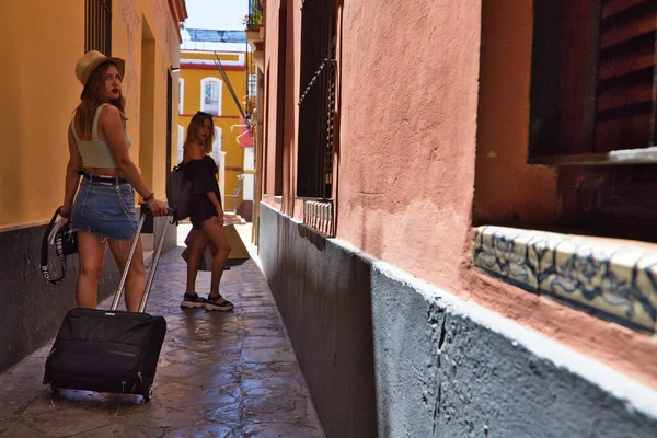 Two beautiful young women walking down a narrow street in a European city. The women are carrying suitcases on wheels and are on their way to the hotel where they are staying for their holidays.