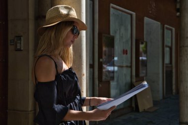 Güneş gözlüklü, şapkalı ve kameralı güzel sarışın kadın ziyaret etmek için şehrin haritasına bakıyor. Turizm, tatil ve seyahat kavramı.