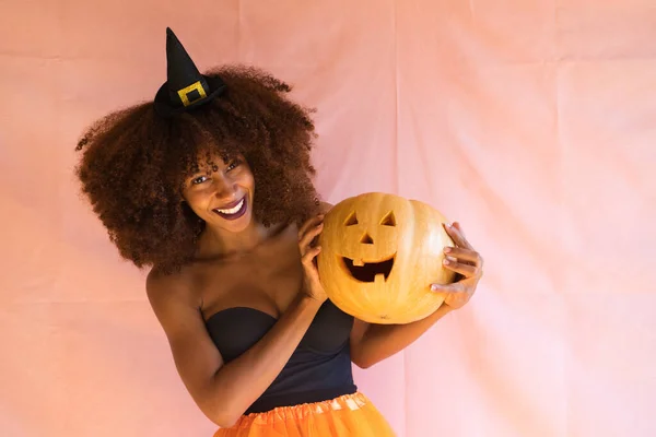 Cadılar bayramı partisi için cadı gibi giyinmiş ve elinde bir balkabağı tutan genç Afrikalı Amerikalı kadın. O bir cadı şapkası ve turuncu bir etek giyer. Kadın kameraya balkabağıyla gülümsüyor..