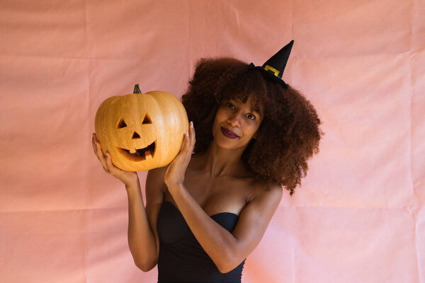 young african american woman dressed as a witch for halloween party and holding a pumpkin in her hands. She wears a witch's hat and an orange skirt. The woman smiles at the camera with the pumpkin.
