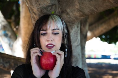 Sarışın, esmer, punky kız elinde kırmızı bir elma tutuyor ve ona bakıyor. Bu kız peri masalından bir cadı..