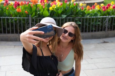 İki genç ve güzel kadın tatilde şehirde seyahat ederken selfie çekiyorlar. Kadınlar kamera, şapka ve güneş gözlüğü takıyor. Tatil, seyahat ve turizm konsepti.