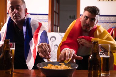 İki erkek arkadaş ve spor hayranı televizyonda bir spor müsabakası izliyorlar. Ellerin kaseden cips kapması gibi detaylar. - Evet. Adamların her biri omuzlarında bir bayrak taşıyor. Spor Konsepti