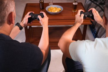 Video konsolunun kumandasını tutan iki arkadaşın elleri hakkında detaylar. Adamlar atıştırmalıklarla bira içerken video oyunları oynuyorlar. Video oyunları ve arkadaşlık kavramı.