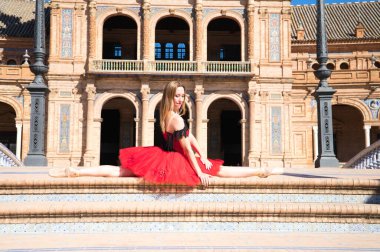 Kırmızı etekli klasik balerin bacaklarını bir parkın merdivenlerine açmış ve kollarını açmış. Klasik bale.