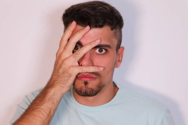 Beyaz arka plandaki genç adam yüzünü tek eliyle açık parmaklarıyla kapatıyor. Adam yüzünde arkadaşsız bir bakışla kameraya bakıyor. İfadeleri kavra