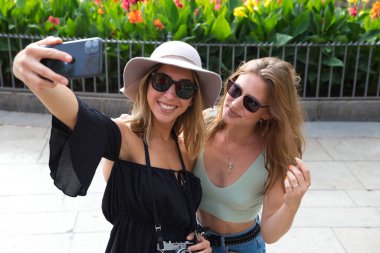 İki genç ve güzel kadın tatilde şehirde seyahat ederken selfie çekiyorlar. Kadınlar kamera, şapka ve güneş gözlüğü takıyor. Tatil, seyahat ve turizm konsepti.