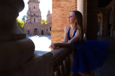 Tavus kuşu mavisi etekli klasik balerin korkuluğa eğilip ufka bakıyor. Balerin güzel ve genç. Klasik bale konsepti.