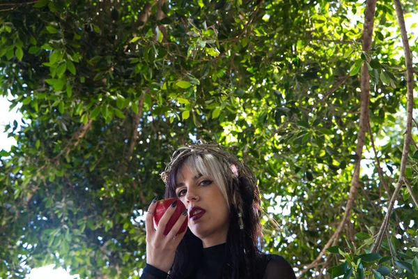 punky girl with blonde and brunette hair holding a red apple in her hands, the girl wears a crown of thorns like christ on her head. The girl is a witch from the fairy tale or the Eve of the present time.