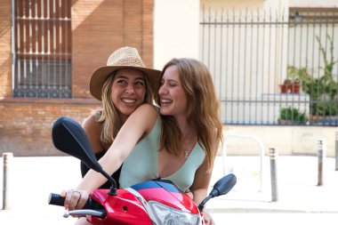 Kırmızı bir motosiklet süren iki genç ve güzel kadın. Kadınlar mutlu ve kameraya ve kendilerine bakarak eğleniyorlar. Kavram Mutluluk, tatil, seyahat eşitliği.