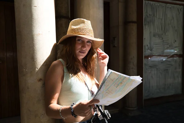 若いと赤い髪のきれいな女の子は 市内の地図を見ている 女の子は帽子をかぶってカメラを持っている 旅行と休日のコンセプト — ストック写真