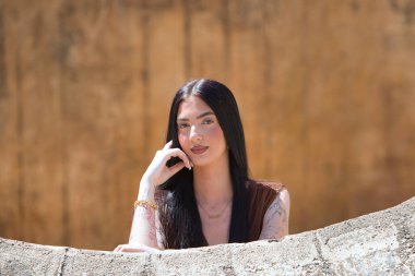 Uzun saçlı, genç, güzel esmer bir kadın yanağını elinde dinlendiriyor ve düşünceli ve bitkin görünüyor. Kadın farklı yönlere bakıyor. Arka planda kahverengi bir duvar var