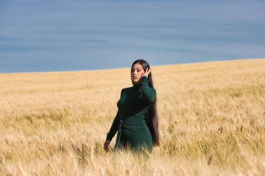 Yeşil giyinmiş, güzel, koyu renk saçlı Latin bir kız yaz günü kuru, sarı bir buğday tarlasının arasında duruyor. Arka planda, mavi gökyüzü