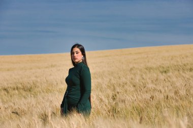Yeşil giyinmiş, güzel, koyu renk saçlı Latin bir kız yaz günü kuru, sarı bir buğday tarlasının arasında duruyor. Arka planda, mavi gökyüzü