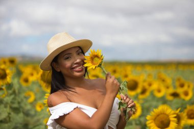 Beyaz gömlek ve hasır şapka giyip ayçiçeklerinin arasında duran koyu tenli genç bir Latin kadın çiftçinin portresi. Kız elinde bir çiçek tutuyor ve kokluyor ve ona bakıyor.