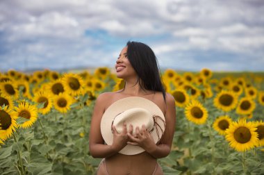 Genç, koyu tenli bir Latin kadın çiftçi, ayçiçeklerinin arasında beyaz bir bikini giyerek duruyor ve göğsünü elinde tuttuğu hasır bir şapkayla örtüyor. Kız doğada eğleniyor.