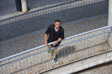 Sakallı, siyah tişörtlü ve bej kargo pantolonlu genç, yakışıklı bir adam, metal bir korkuluğa tutunuyor, rampadan aşağı indi ve yukarı bakıyor. Fotoğraf yukarıdan çekildi