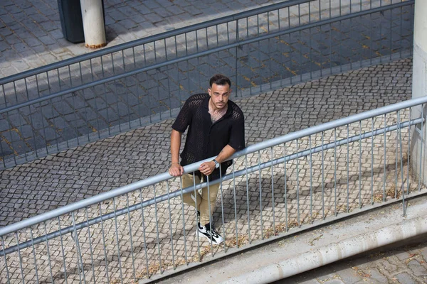 Sakallı, siyah tişörtlü ve bej kargo pantolonlu genç, yakışıklı bir adam, metal bir korkuluğa tutunuyor, rampadan aşağı indi ve yukarı bakıyor. Fotoğraf yukarıdan çekildi