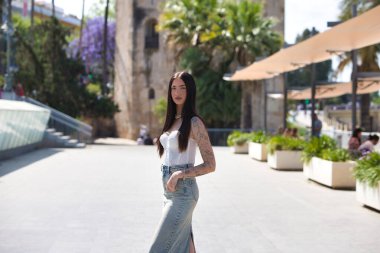 Kollarında dövmeleri olan, sıradan kıyafetler giyen, güneşli bir günde şehrin büyük bir caddesinde poz veren genç ve güzel bir Latin kadın. Arka planda Sevilla 'nın altın kulesi var.