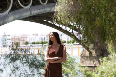 Uzun kahverengi saçlı, kahverengi elbiseli ve siyah çizmeli çekici bir İspanyol kadın Sevilla 'daki Triana Köprüsü' nün yanındaki nehir kıyısında duruyor. Kadın güvenle kollarını kavuşturuyor.