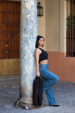 Genç, çekici, esmer, kot pantolon giyen ve siyah bluzlu bir kadın elinde bir blazer ceket tutuyor ve İspanya 'nın merkez bölgesinde bir caddede mermer bir sütuna yaslanıyor..