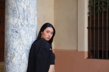Ciddi, genç ve çekici bir kadının portresi, koyu renk saçlı, siyah bluzlu, ceketini omzunun üstünde tutan ve Seville 'in merkezindeki mermer sütuna yaslanan.