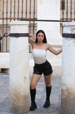 Beyaz dantelli bluz giyen, siyah şortlu ve deri çizmeli genç, çekici bir İspanyol kadın Seville Katedrali 'nin yanındaki iki taş sütun arasında çeşitli pozlar veriyor.