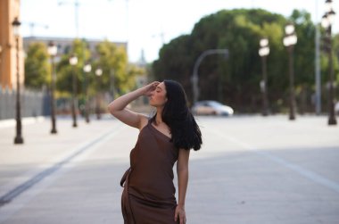 Kahverengi elbiseli genç, çekici bir İspanyol kadın Sevilla, İspanya 'da bir caddede manken gibi yürüyor. Kadın yürürken poz veriyor. Konsept: güzellik ve moda