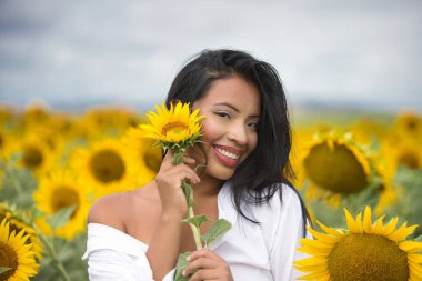 Beyaz gömlekli güzel bir melez Latin kadın onu yüzüne yaklaştırıyor ve bir ayçiçeğiyle flört ediyor. Kadın ayçiçeklerinin arasında mutlu ve güneşli yaz gününde gülümsüyor.