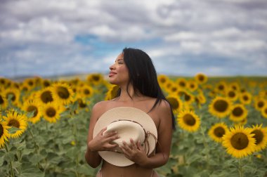 Genç, koyu tenli bir Latin kadın çiftçi, ayçiçeklerinin arasında beyaz bir bikini giyerek duruyor ve göğsünü elinde tuttuğu hasır bir şapkayla örtüyor. Kız doğada eğleniyor.
