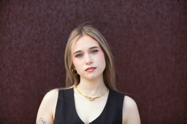 Açık renk gözlü sarışın kadın portresi, siyah giyinmiş genç ve güzel paslı metal bir arka planda kameraya bakıyor.